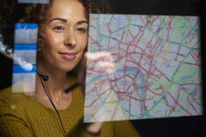 A young woman with a curious expression stands in front of a map, attentively examining it with her finger tracing a route. She appears engaged and focused, ready to explore and navigate the world